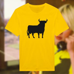 Puede incluir: Una camiseta amarilla con una silueta negra de un toro en la parte delantera.