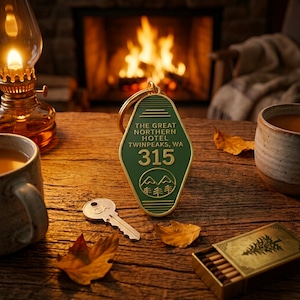 May include: A dark green, diamond-shaped motel-style keyring with gold-coloured trim and lettering. The text reads THE GREAT NORTHERN HOTEL TWINPEAKS, WA 315, accompanied by a graphic of mountains and pine trees. A metal key is positioned nearby.