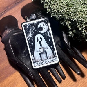 May include: A black and white tarot card featuring a ghost holding a cup of coffee. The card is titled "The Coffee Lovers".