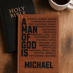 Puede incluir: Un diario de aspecto de cuero marrón con el texto negro impreso A MAN OF GOD IS seguido de una lista de virtudes que incluyen faithful, humble, strong, courageous y el nombre MICHAEL en la parte inferior, junto a una Santa Biblia negra.
