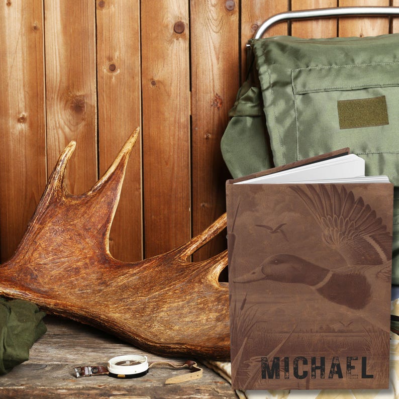 A brown personalized hunting journal with a duck design and the name “Michael” sits propped open against a rustic wooden backdrop. A large antler and green outdoor backpack rest nearby, with a compass and leather strap placed on a weathered wooden surface. The earthy colors and hunting gear emphasize a rugged outdoor theme, highlighting the journal as a keepsake for hunters and nature lovers.