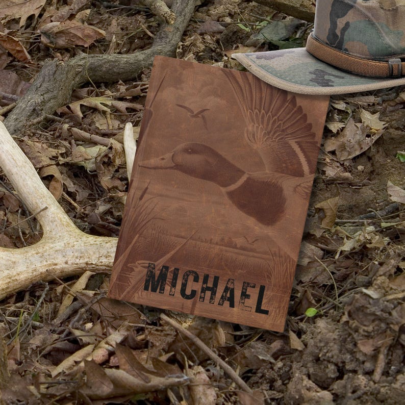 The personalized hunting journal with a duck illustration and the name “Michael” lies on the forest floor among dried leaves, branches, and a deer antler. A camouflage hunting cap rests nearby, reinforcing the outdoorsman theme. The earthy tones of the journal blend seamlessly with the woodland setting, presenting the book as a rugged and masculine keepsake for hunters or nature enthusiasts to document outdoor adventures.