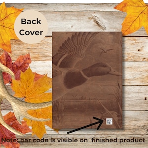 The back cover of a brown duck hunting journal is displayed against a rustic wooden surface surrounded by colorful autumn leaves and a deer antler. The cover features the same engraved duck design as the front, and a white barcode is visible at the bottom right corner with a black arrow pointing to it. A tan circle reads “Back Cover,” noting that the barcode will remain on the finished product.
