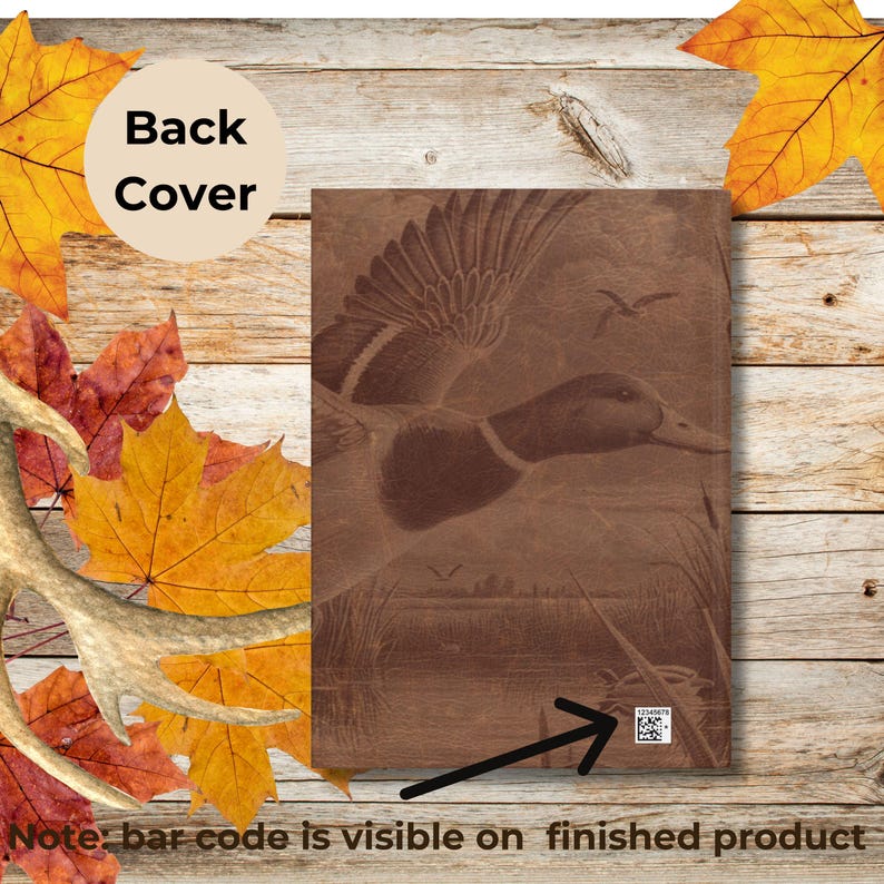 The back cover of a brown duck hunting journal is displayed against a rustic wooden surface surrounded by colorful autumn leaves and a deer antler. The cover features the same engraved duck design as the front, and a white barcode is visible at the bottom right corner with a black arrow pointing to it. A tan circle reads “Back Cover,” noting that the barcode will remain on the finished product.
