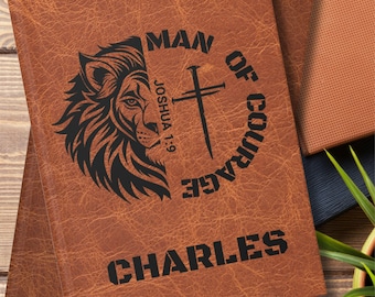 Può includere: Diario in pelle marrone con un disegno a testa di leone e croce con le parole "MAN OF COURAGE". Il nome "CHARLES" è stampato in basso. È visibile anche il testo "Joshua 1:9".