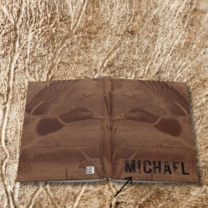 An open view of the personalized hunting journal shows the full cover spread laid flat against a tan textured background resembling tree bark. The cover displays two mirrored flying ducks with wings extended, cattails at the bottom, and the name “Michael” printed boldly across the lower right. A small barcode appears at the bottom left corner, highlighted by a black arrow. The design emphasizes the wraparound artwork and natural theme.
