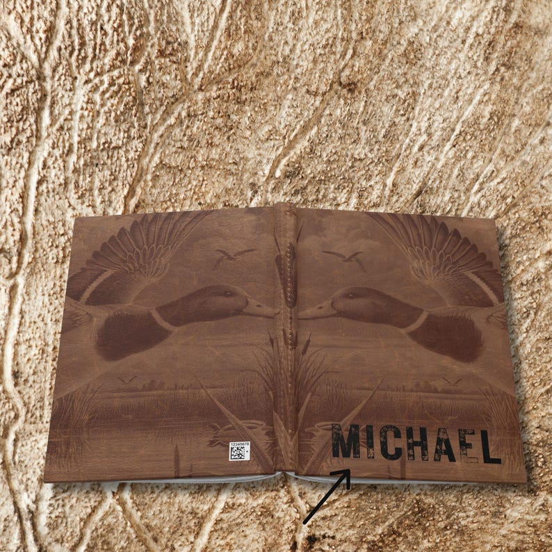 An open view of the personalized hunting journal shows the full cover spread laid flat against a tan textured background resembling tree bark. The cover displays two mirrored flying ducks with wings extended, cattails at the bottom, and the name “Michael” printed boldly across the lower right. A small barcode appears at the bottom left corner, highlighted by a black arrow. The design emphasizes the wraparound artwork and natural theme.