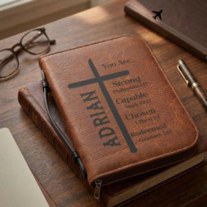 May include: A brown leather Bible cover featuring the name "ADRIAN" and a cross. The cover is inscribed with "You Are..." followed by "Strong," "Capable," "Chosen," and "Redeemed" with associated Bible verses. A zip closure is visible.