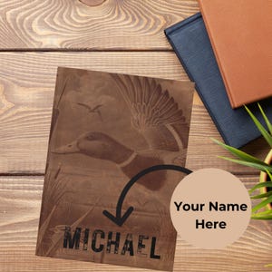 A personalized hunting journal with a duck engraving and the name “Michael” is placed on a wooden desk next to stacked black and brown textured books and a green plant in a yellow pot. A bold black arrow points to the customizable name section with a tan circle reading “Your Name Here.” The rustic wood surface and natural accents emphasize personalization and make the journal ideal as a custom men’s gift.