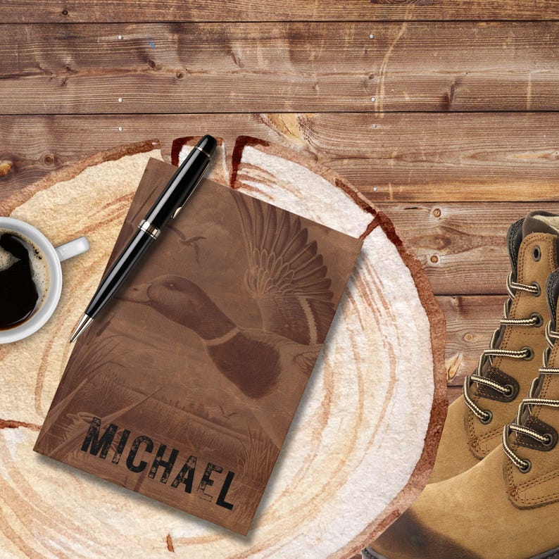 A personalized hunting journal featuring a flying duck design and the name “Michael” rests on a round wooden slab with visible growth rings. A sleek black pen lies across the journal, while a steaming cup of black coffee sits on the table beside rugged tan hunting boots. The wood plank backdrop and outdoor-inspired details create a masculine, rustic atmosphere, showing the journal as perfect for hunters or outdoorsmen.