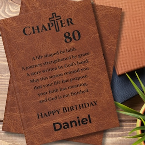 May include: A brown faux leather journal with a textured surface featuring a black cross icon and the text: CHAPTER 80. A life shaped by faith. A journey strengthened by grace. A story written by God's hand. May this season remind you that your life has purpose, your faith has meaning, and God is not finished. HAPPY BIRTHDAY Daniel.