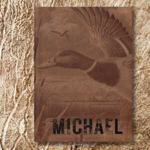 A close-up of the personalized duck hunting journal rests upright against a bark-like tan textured background. The name “Michael” is boldly printed across the bottom in distressed black font, while the cover art depicts a flying duck with wings outstretched above a marsh landscape. The earthy wood-grain tones of the journal blend with the background, creating a rugged, natural presentation that emphasizes outdoor adventure and rustic style.