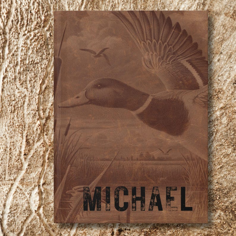 A close-up of the personalized duck hunting journal rests upright against a bark-like tan textured background. The name “Michael” is boldly printed across the bottom in distressed black font, while the cover art depicts a flying duck with wings outstretched above a marsh landscape. The earthy wood-grain tones of the journal blend with the background, creating a rugged, natural presentation that emphasizes outdoor adventure and rustic style.