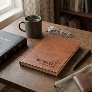 May include: A brown leather journal with the name "MICHAEL" and a cross symbol, along with the text "philippians 4:13". A black pen, a cup of coffee, and a pair of glasses rest on a wooden table. A black Bible is also present.