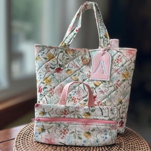 May include: A quilted tote bag and a matching zippered pouch featuring a floral pattern with yellow, pink, and green botanical elements. The tote includes a wooden button closure and a pink rectangular luggage tag embroidered with the letter A.