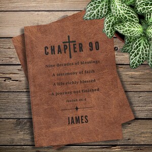 Könnte beinhalten: Ein brauner Bucheinband in Lederoptik mit einem schwarzen Kreuzsymbol und dem Text: CHAPTER 90, Nine decades of blessings, A testimony of faith, A life richly blessed, A journey not finished, Isaiah 46:4, sowie dem Namen JAMES.