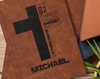 Può includere: Diario rilegato in pelle marrone con un design a croce nera e il testo "I CAN DO ALL THINGS THROUGH CHRIST WHO STRENGTHENS ME" e il nome "MICHAEL". Il diario è impilato con altri diari.