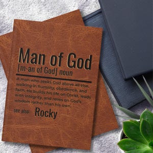 以下が含まれることがあります： 「Man of God」の文字が書かれた茶色の革製ジャーナル。 「Man of God」の定義がタイトルの下に印刷されています。 ジャーナルは、他の本や植物と一緒に、テクスチャ加工された表面に置かれています。