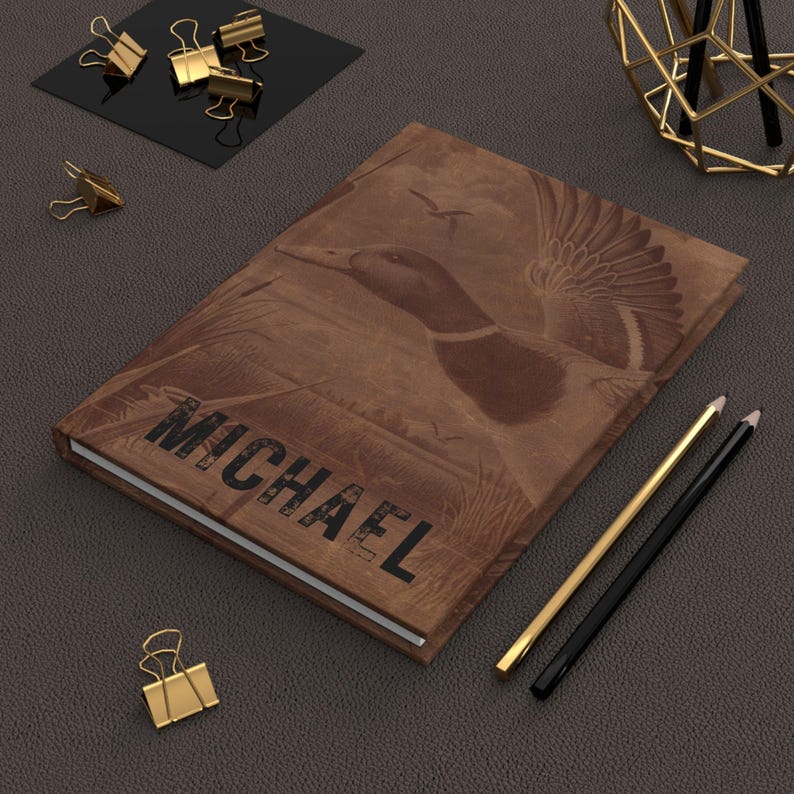 A brown hunting-themed journal with the name “Michael” engraved at the bottom lies on a textured dark gray leather surface. The cover shows a detailed duck in flight above cattails. Around it are golden binder clips, a modern black-and-gold wire pencil holder, and gold and black pencils. A white arrow points toward the customizable name. The refined, professional setup contrasts with the rustic design, making it appealing for everyday use.
