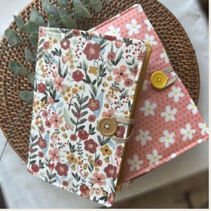 May include: Two rectangular fabric book covers with quilted stitching. The front cover features a white background with a multi-colored floral pattern and a tan button closure. The second cover is pink with a white daisy pattern and a yellow button.
