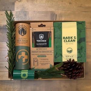 May include: A gift box containing a 2 oz Rhino Wax healing paw balm tube, a TickCheck premium tick remover set with tools and pouch, and a pack of Bark & Clean pet wipes measuring 8.5 inches by 9 inches, accented with a pine branch and pinecone.