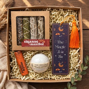 May include: A gift box containing a Cleanse + Manifest smudge stick set, a dark purple bookmark with an orange tassel and the text The Magic Is in You, a round white bath bomb labeled The Dreamer, and an orange tube labeled Poppy & Pout.