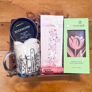May include: A gift box with a mug, candle, bookmark, lip balm, and chocolate bar. The mug has a book design. The candle is labeled "BOOKWORM". The bookmark has a floral design. The chocolate bar is labeled "Spring Tulip".