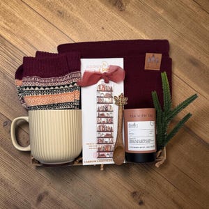 May include: A gift set containing a cream-colored ribbed mug, a pair of maroon patterned socks, a maroon beanie with a leather patch, a bookmark with a bookshelf illustration, a wooden spoon with a leaf handle, and a tea tin labeled Tea With Tae.