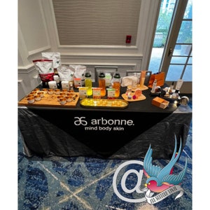 May include: A black table covered with a black tablecloth with the words "arbonne. mind body skin" printed in white. The table is set with a variety of products, including bottles of liquid, small containers, and bowls. The products are arranged in a symmetrical pattern.