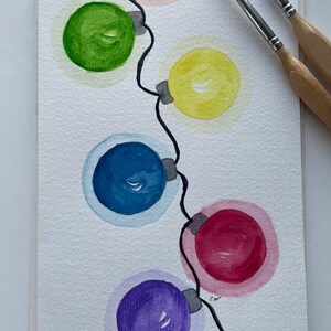 May include: Watercolor painting of a string of Christmas lights in various colors: orange, green, yellow, blue, red, and purple. The lights are connected by a black wire. Two paintbrushes are in the upper right corner. The artwork is on a white background.