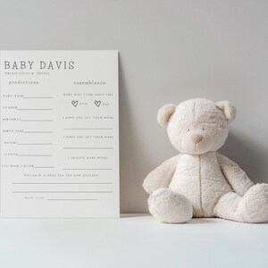 May include: A cream teddy bear sits beside a white baby prediction card. The card reads "Baby Davis" and has spaces for predictions about the baby's name, gender, and other details. The bear has a soft, plush appearance.