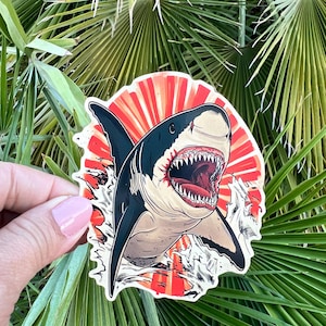 May include: A sticker featuring a detailed illustration of a shark with an open mouth, revealing sharp teeth. The shark is depicted in shades of gray, black, and beige, set against a red and white background. The sticker is held against a backdrop of green palm leaves.