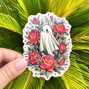 May include: A sticker featuring a friendly ghost surrounded by red roses and green leaves. The ghost is white with a simple face. The sticker has a textured, speckled background.