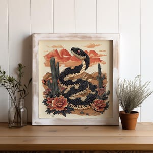 Könnte beinhalten: Gerahmter Kunstdruck mit einer stilisierten Darstellung einer Schlange in einer Wüstenlandschaft. Die Schlange ist schwarz und cremefarben mit roten Augen, umgeben von Kakteen, Blumen und Bergen. Der Rahmen ist weiß mit einem Used-Look.