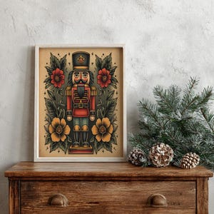 Può includere: Stampa incorniciata con un'illustrazione in stile vintage di un soldato schiaccianoci. L'opera presenta lo schiaccianoci in uniforme rossa, circondato da fiori rossi e gialli e fogliame verde, su sfondo beige. La stampa è su una cassettiera di legno.