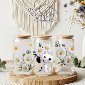 May include: Three glass tumblers with bamboo lids and clear straws. Each tumbler features a daisy design, with one showcasing a Snoopy character. The tumblers are displayed on a wooden tray, with a vase of flowers in the background.