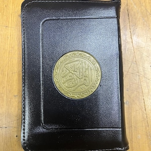 May include: A black leather-bound book with a gold zipper closure and a gold circular emblem featuring Arabic calligraphy.