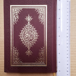 May include: A burgundy book with gold embossed lettering and decorative patterns. The cover features a central medallion and ornate border. A ruler is placed next to the book, showing a length of approximately 20 cm.