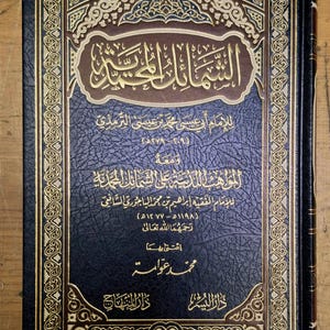 Puede incluir: Un libro de tapa dura azul oscuro con diseños dorados y marrones. La portada presenta texto árabe dorado, con intrincados patrones de borde. El libro parece ser un texto religioso.