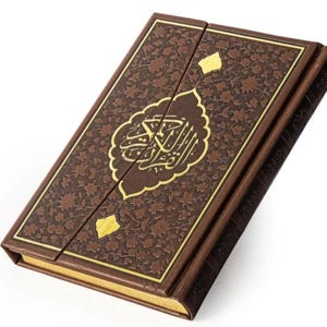 May include: A brown leather-bound book with gold accents and ornate floral patterns. The cover features gold Arabic calligraphy within a decorative frame. The book is closed, with a magnetic clasp.