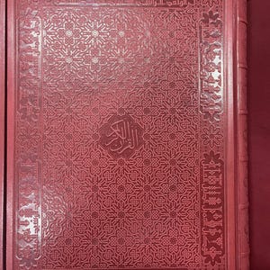 Op de afbeelding: Een rood boek met ingewikkelde reliëfpatronen en Arabisch schrift. De omslag heeft een gedetailleerd geometrisch ontwerp en een centraal embleem. Het boek zit in een bijpassende rode hoes.