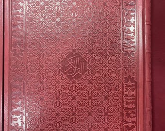 May include: A red book with intricate embossed patterns and Arabic script. The cover features a detailed geometric design and a central emblem. The book is in a matching red case, suggesting a high-quality edition.