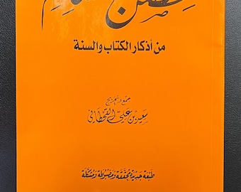 May include: An orange book with Arabic script on the cover. The title, in large black calligraphy, reads "Hisn al-Muslim." Below the title are additional lines of text in Arabic. The book's cover also includes the author's name and publisher's logo.