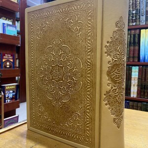 May include: A tan leather-bound book with intricate gold embossed designs and Arabic script. The book is closed and standing upright on a wooden surface.