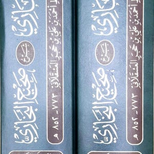 May include: Two hardcover books with dark teal covers. The covers feature ornate gold and white Arabic script and decorative patterns. The spines display text in Arabic. The books are part of a series.