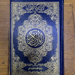 May include: A navy blue book with gold ornate designs and Arabic script. The cover features a central medallion and decorative borders. The book is closed and appears to be in good condition.