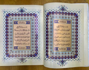 May include: Open book with two pages featuring ornate calligraphy. The pages have a cream-colored background with intricate blue, gold, and red decorative borders. The text is in Arabic script, with each page containing multiple lines of text.