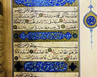 Könnte beinhalten: Seite aus einem religiösen Buch mit arabischer Kalligraphie. Der Text ist in schwarzer Tinte, mit dekorativen blauen und goldenen Bordüren. Die Seite hat einen cremefarbenen Hintergrund und stammt aus einem offenen Buch.