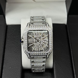 May include: A silver wristwatch with a square face and a skeleton dial. The watch is adorned with many small, clear gemstones on the bezel and strap. The watch is presented in a black box with a white cushion.