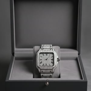 May include: A silver wristwatch with a square face and a matching band, encrusted with small, sparkling stones. The watch is displayed inside a gray box with a hinged lid, set against a gray background. Roman numerals mark the hours on the watch face.